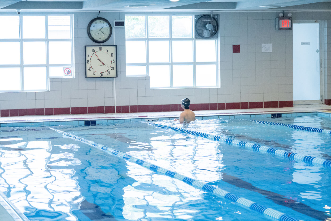 Swimmer resting at the edge of an indoor lap pool with lane dividers, wall clocks, bright windows, and an EXIT sign.