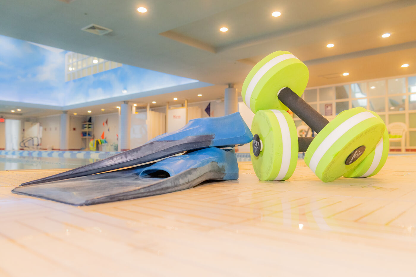 Bright indoor pool deck with blue swim fins and neon green water dumbbells on the floor, lit by warm ceiling lights.