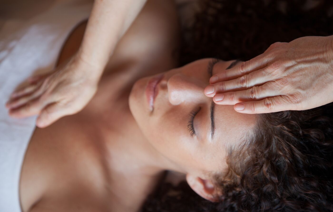 A woman with closed eyes receives a forehead massage from a practitioner’s hands. Relaxing mood, neutral background, and soft lighting.
